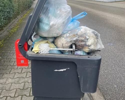 Das Foto zeigt eine überfüllte graue Mülltonne am Straßenrand. Die Tonne kann nicht geschlossen werden, weil sich Müllsäcke darauf stapeln. Foto MERB, Achern