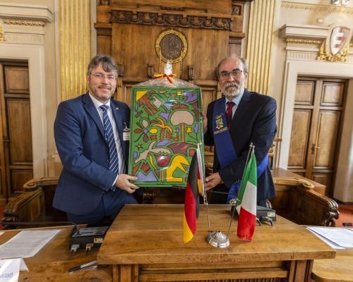 Das Foto zeigt Landrat Prof. Dr. Christian Dusch (links) und Giuseppe Paolini, Präsident der Provinz Pesaro-Urbino, hinter einem Holztisch, auf dem eine deutsche und eine italienische Flagge stehen. Beide halten ein Geschenk in der Hand.