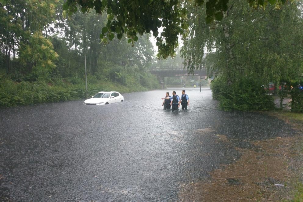 Das Foto zeigt eine überschwemmte Straßes mit drei Personen und einem tief im Wasser fahrenden Auto.