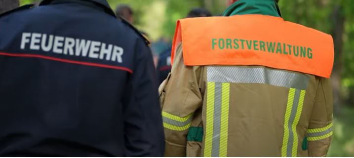 Zwei Personen sind mit dem Rücjen zu sehen. Auf ihren Arbeitsuniformen steht Feuerwehr und Forstverwaltung.